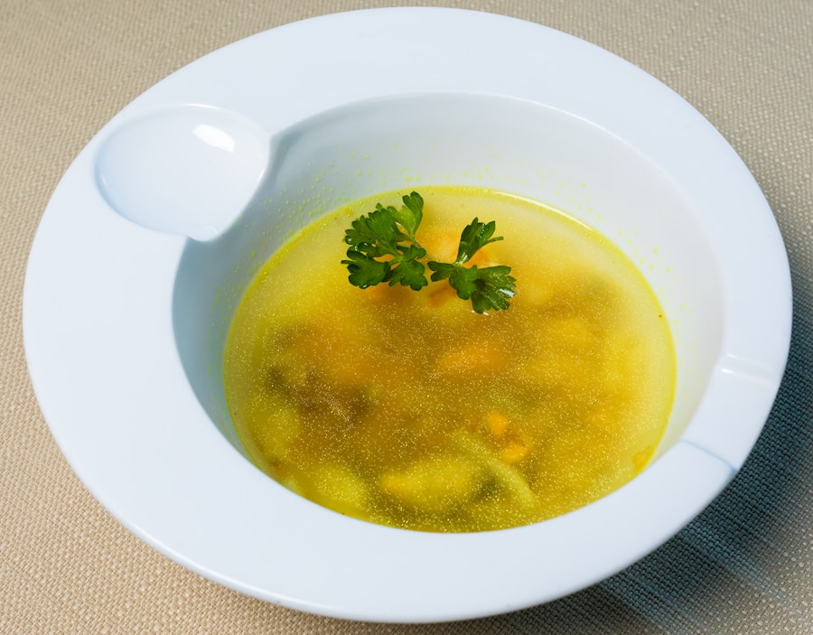 Eine weiße Schale, gefüllt mit klarer gelber Suppe mit kleinen Gemüsestückchen, garniert mit zwei Zweigen frischer Petersilie, steht auf einer strukturierten beigen Fläche.