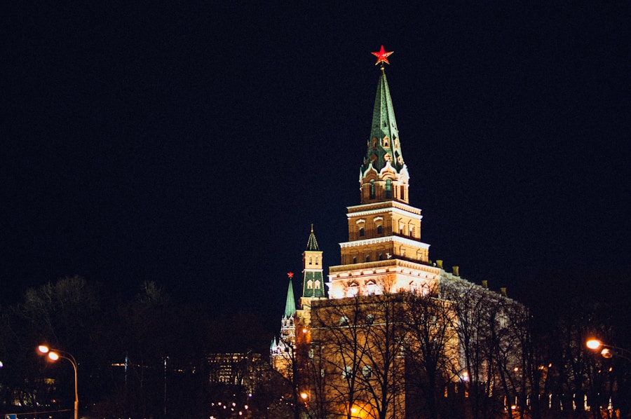 Ein hoher, beleuchteter Kremlturm mit einer grünen Spitze und einem roten Stern auf der Spitze steht am Nachthimmel, umgeben von Bäumen und Lichtern der Stadt.